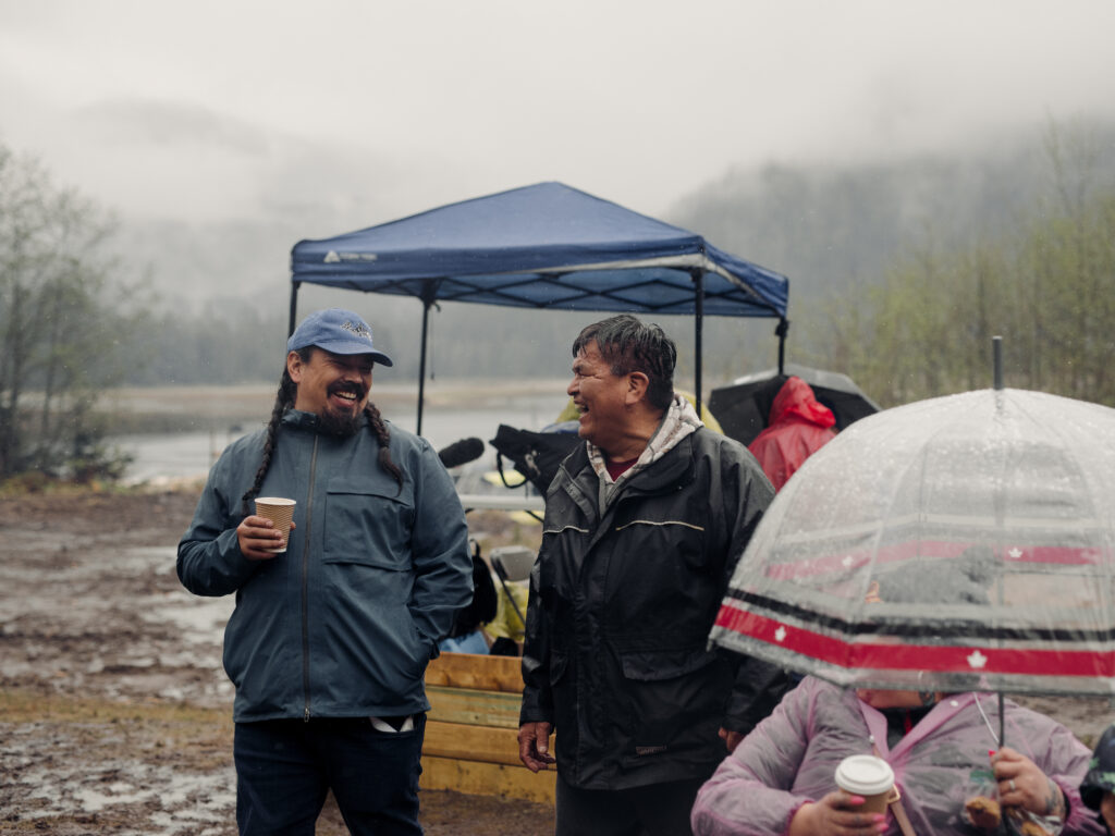 Two Mamalilikulla men laugh, one holding a cup of coffee, wearing rain jackets. A person with an umbrella is in the foreground.
