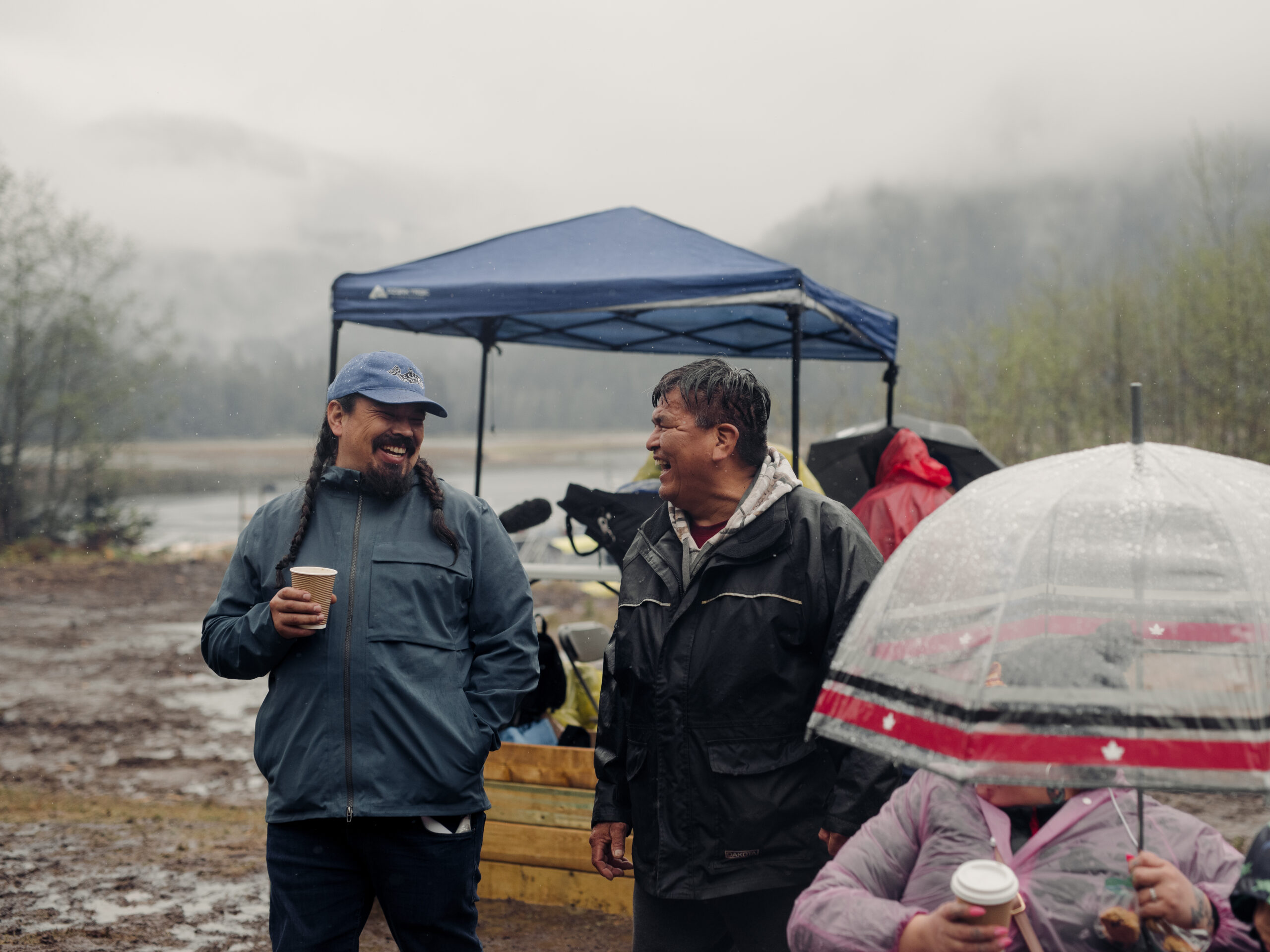 Two Mamalilikulla men laugh, one holding a cup of coffee, wearing rain jackets. A person with an umbrella is in the foreground.