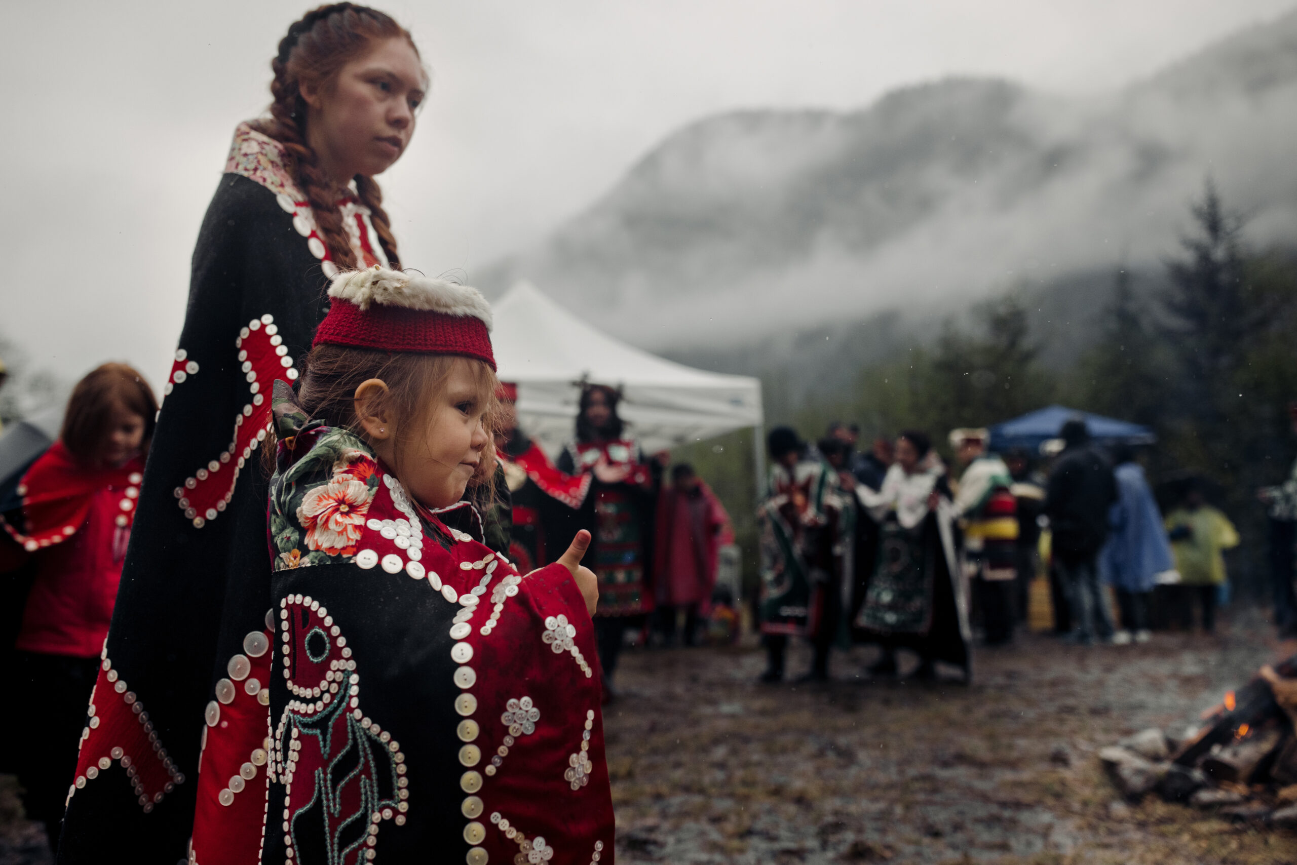 A small child and Mamalilikulla youth dance in regalia at Mamalilikulla's recently proclaimed protected area. People watch in the background, and misty mountains are visible