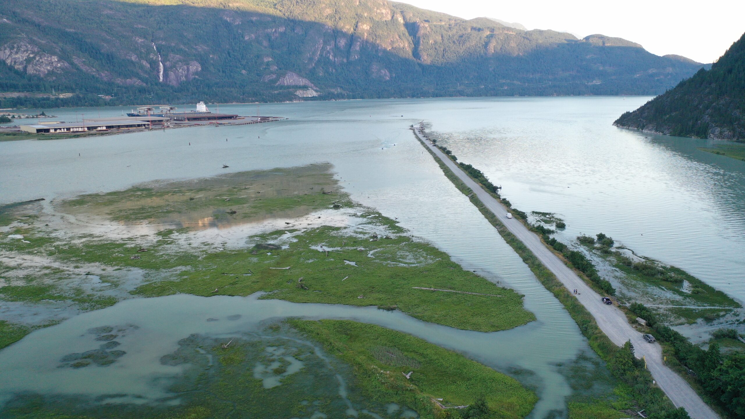 Squamish River spit: An aerial few of a 5-kilometre spit stretching into the ocean water of Howe Sound. On the right is the Squamish River mouth. On the left is green marshy waters, which is the Squamish Estuary. Mountains are visible in the background.