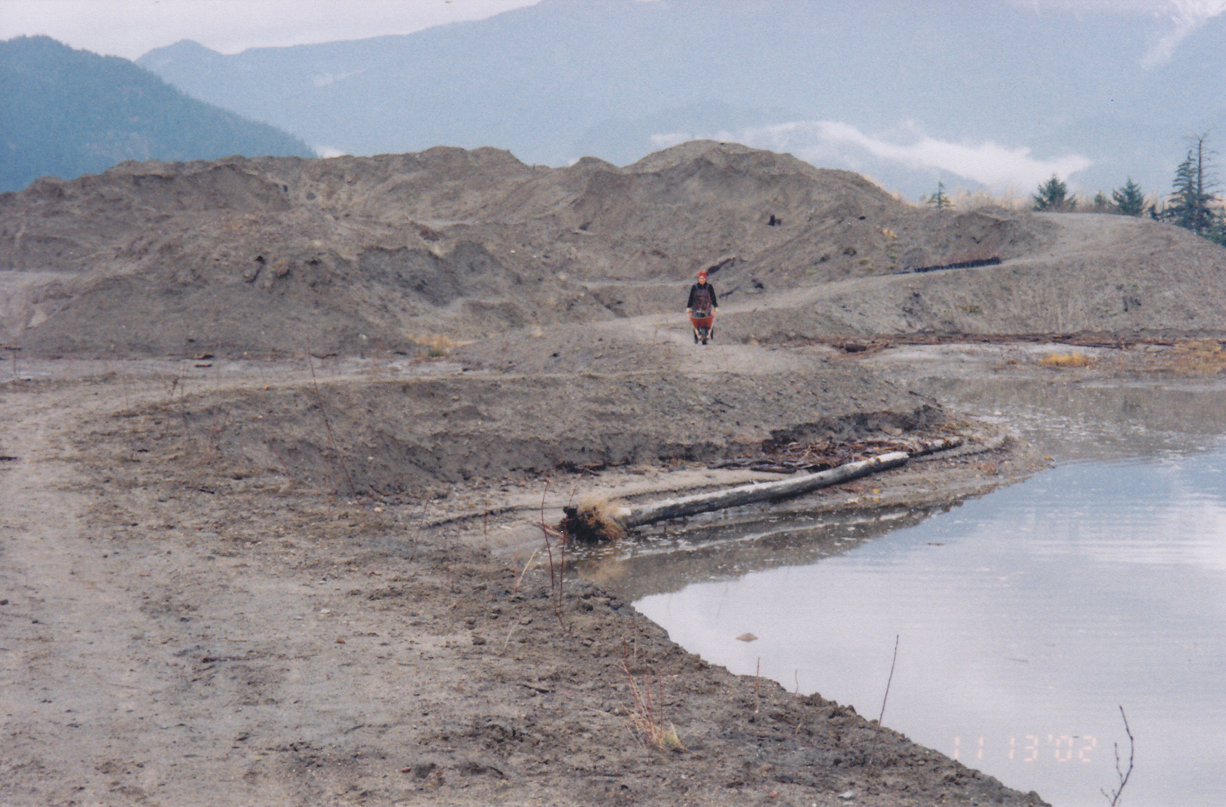 Squamish estuary: A photo of the Squamish estuary before undergoing restoration, showing a barren, brown dirt landscape with mountains in the background. A long figure is visible biking on a dirt path. A fallen log and large puddle are in the foreground.