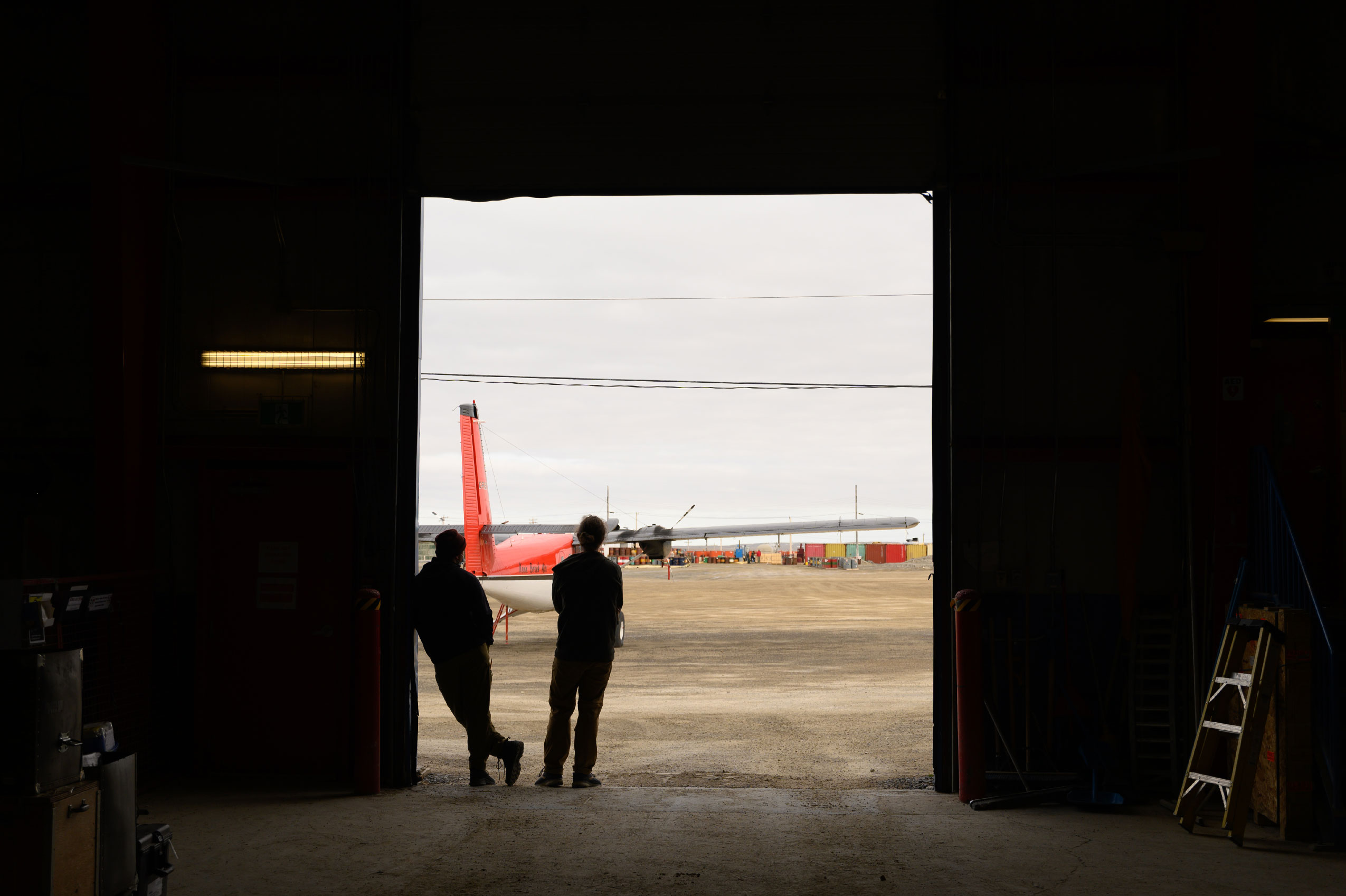 View from inside a building with 2 people standing in the large doorway looking outside at an airplane at the the Polar Continental Shelf Program Base. 