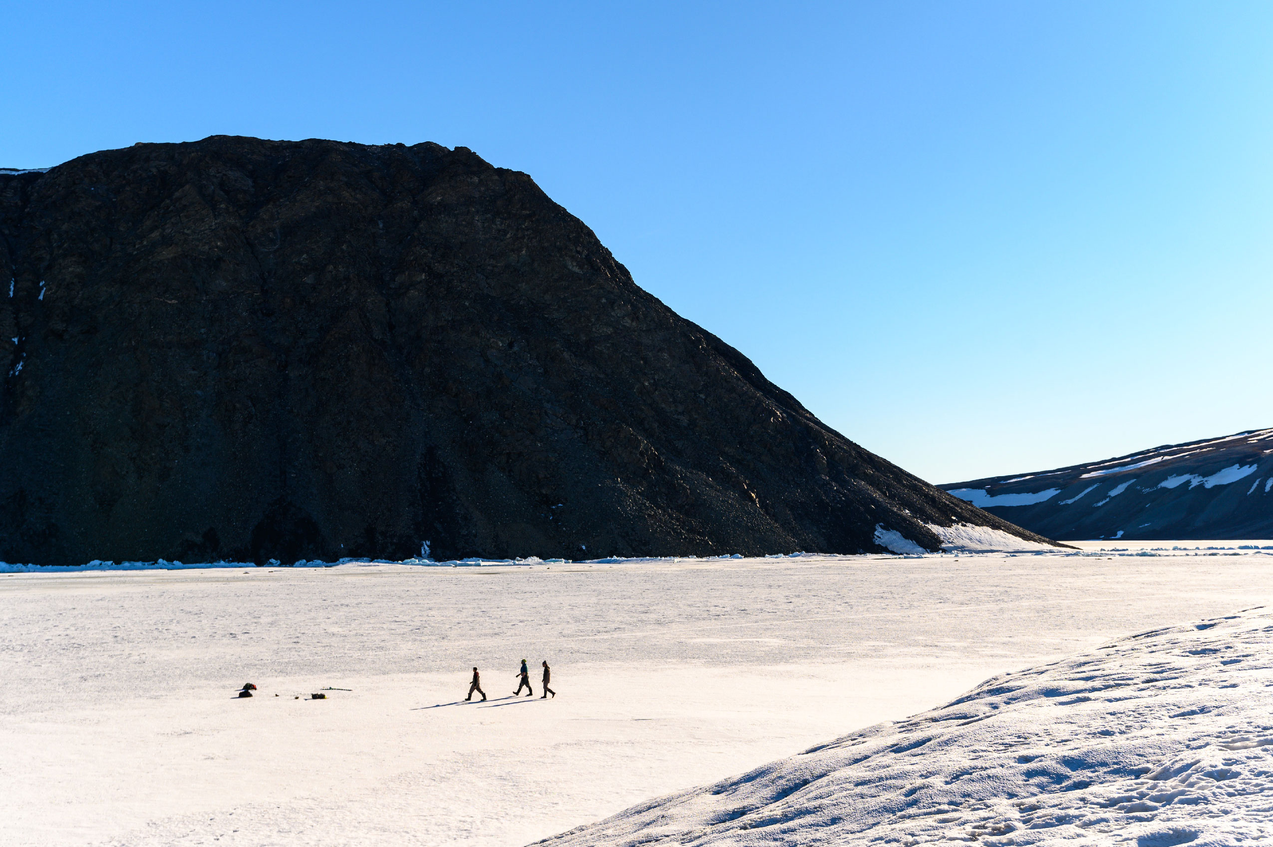 Three people walking on ice shelf and the glacial tongue in the foreground with a large rock formation behind them.