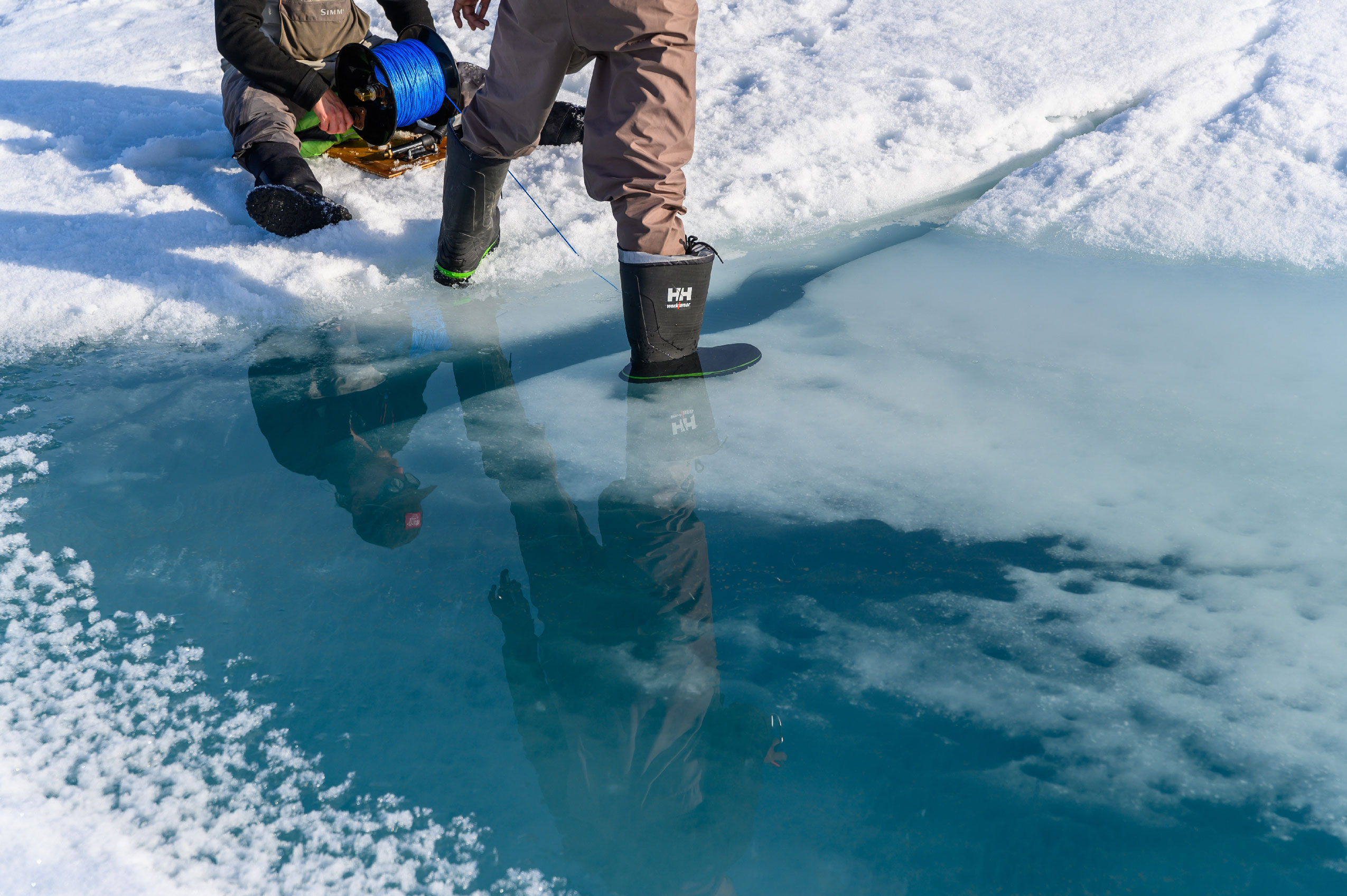 Two scientists on the ice with a long cable on a spindle. One scientist has a foot submerged in water halfway up their boot.