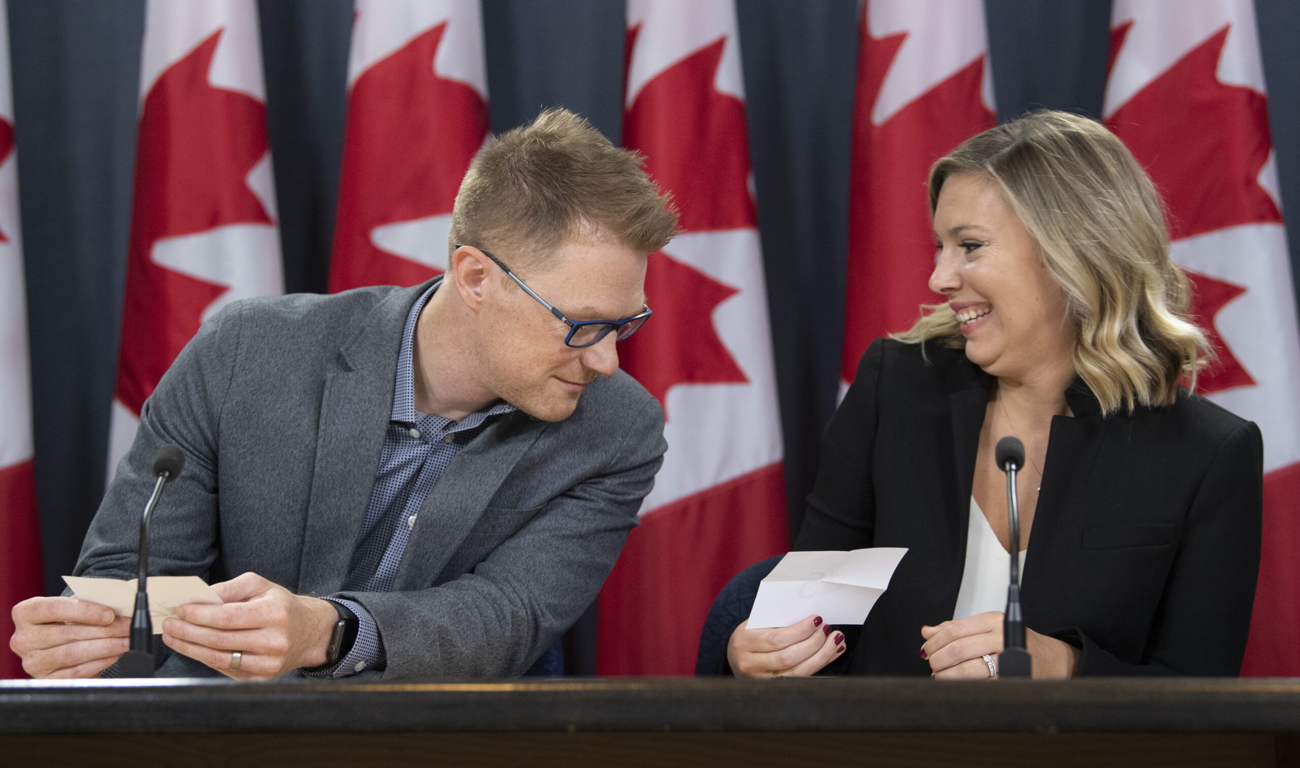 A man and a woman are sitting at a desk with Canadian flags in the background. They are both holding small pieces of white paper. The man, Brock Harrison, is wearing a grey-blue blazer with blue button shirt, blue-rimmed glasses and short-cropped hair. He is looking over at the woman,s paper. The woman, Kate Purchase, is wearing a black blazer with a white shirt, and shoulder-length hair. She is smiling.