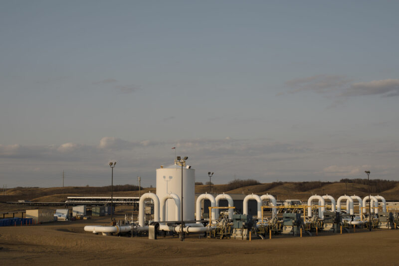 A series of white painted pipes and other oil and gas infrastructure is set against a landscape of brownish ground and dull blue sky.