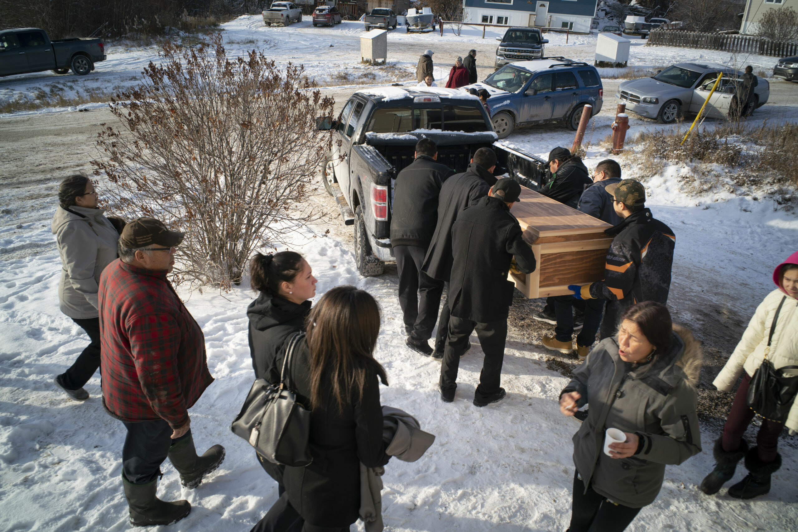A casket being loaded onto a truck with people gathering nearby on a wintry day