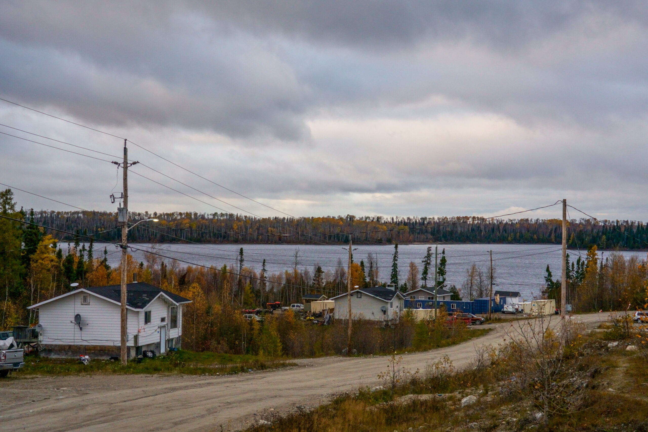 A dirt road leading past houses and power lines and a lake, with autumn colours on the trees