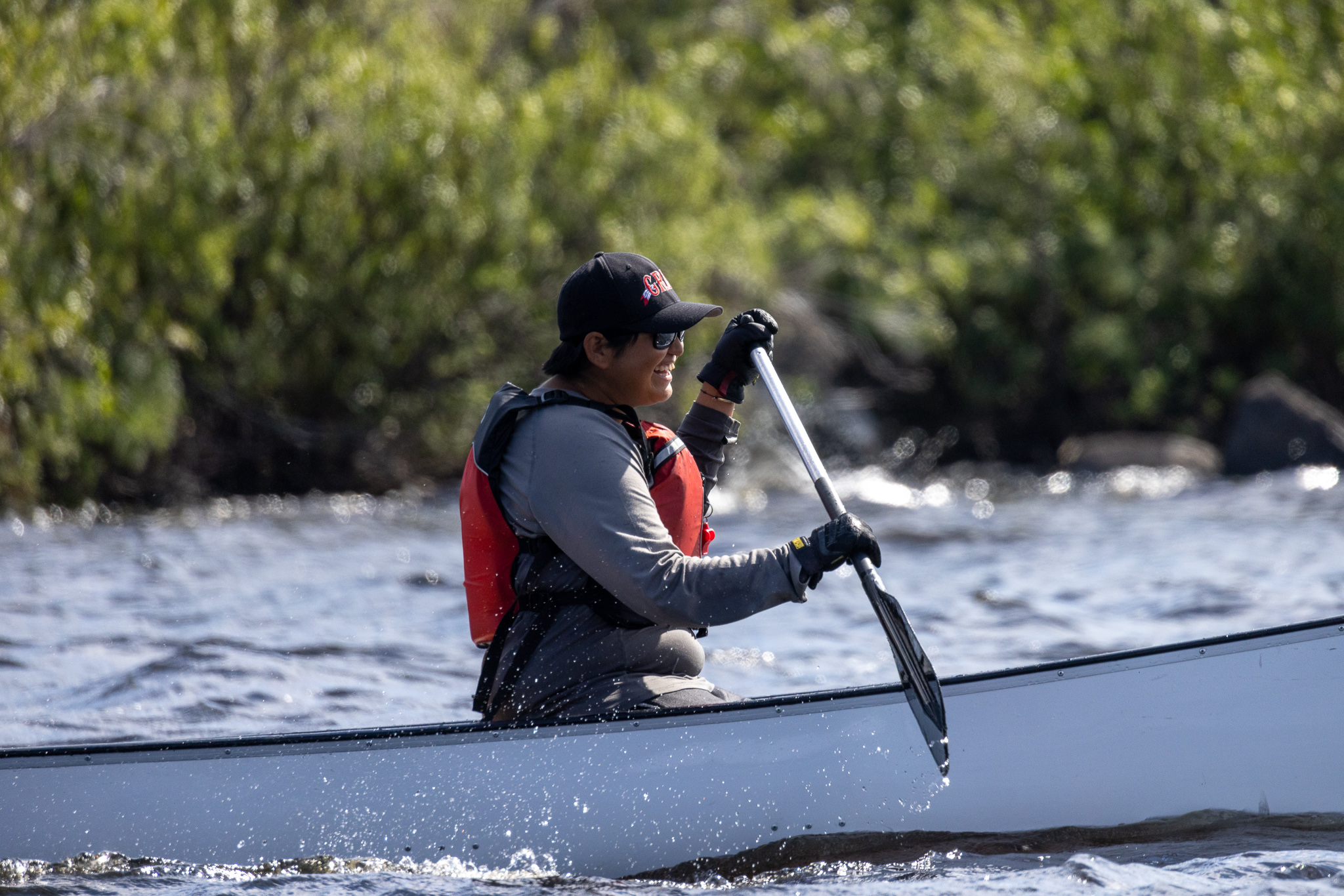 Martina Denechezhe smiles as she paddles a grey canoe wearing a black cap and red life vest