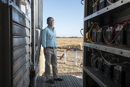 A man in a blue shirt and grey pants, David Tian, stands at the doorway of a structure with computers lining the walls. A grain field can be seen outside and a gas pipeline.