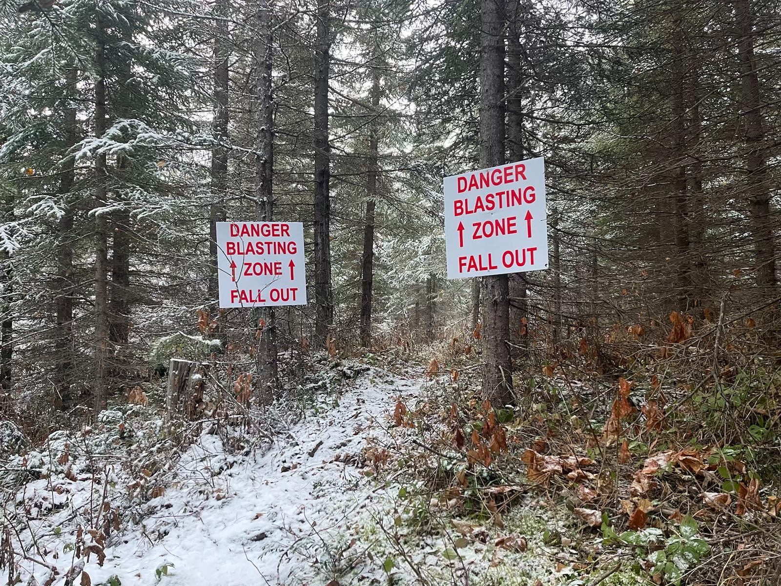 Snowy forested trail with signs on trees noting "danger blasting zone fall out"