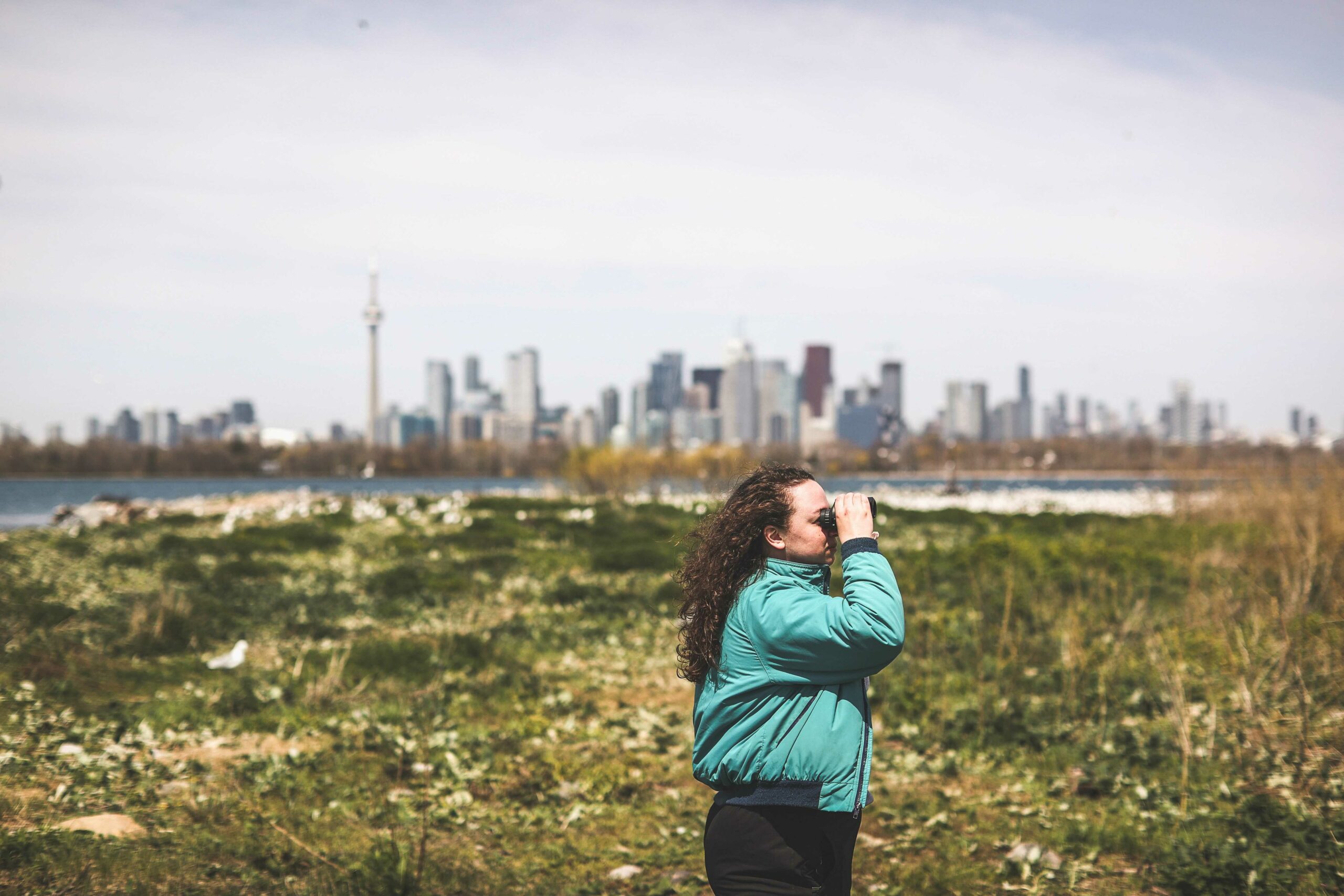 Reporter Emma McIntosh looking into the distance at Tommy Thompson Park using binoculars. 