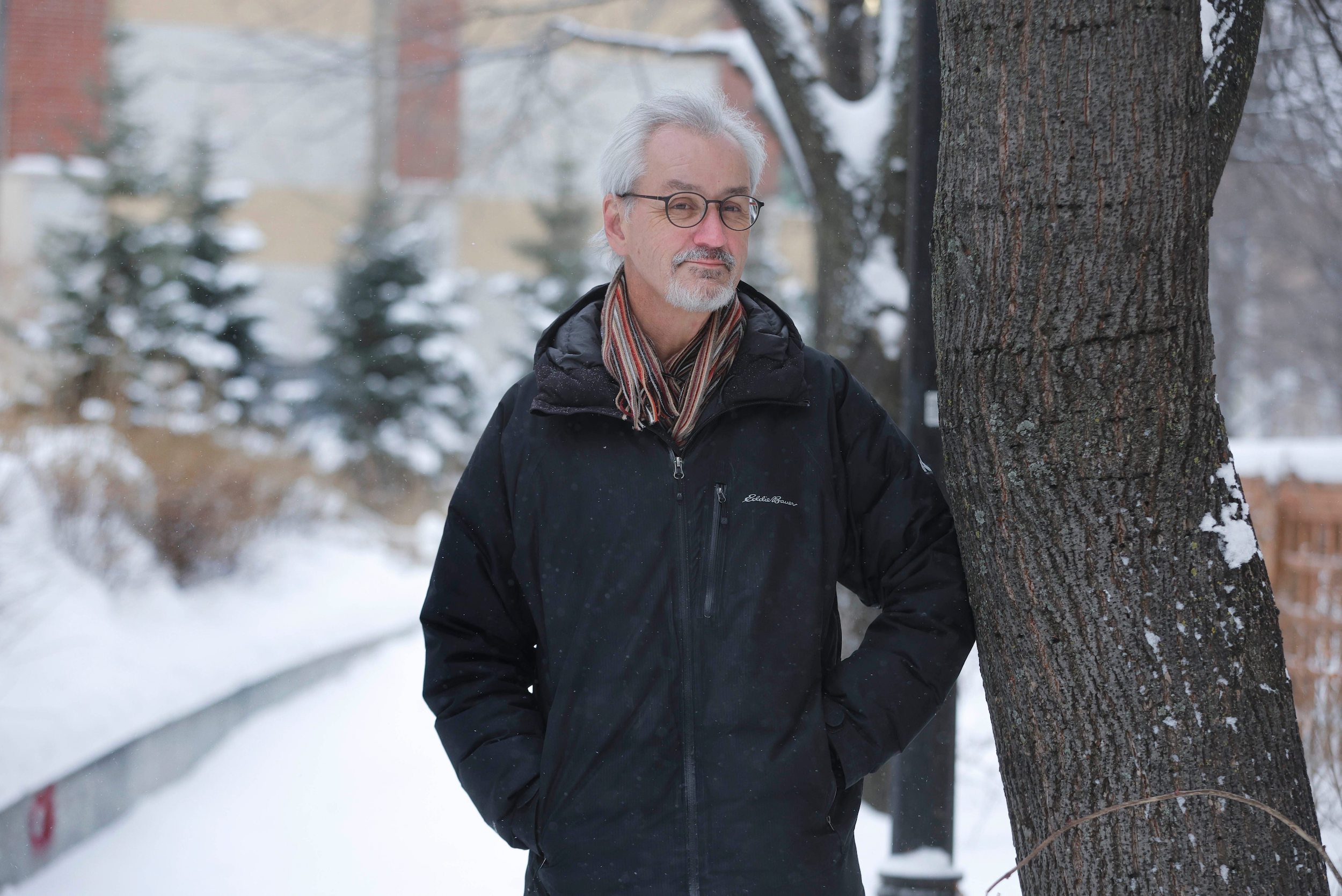 Landscape architect Glen Manning leans against a tree on a winter day in Winnipeg's downtown exchange district. He wears a dark jacket and striped scarf
