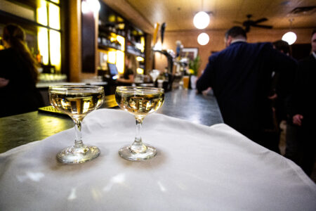 Two glasses of white wine sit on a white tablecloth at a restaurant with people in suits in the background.
