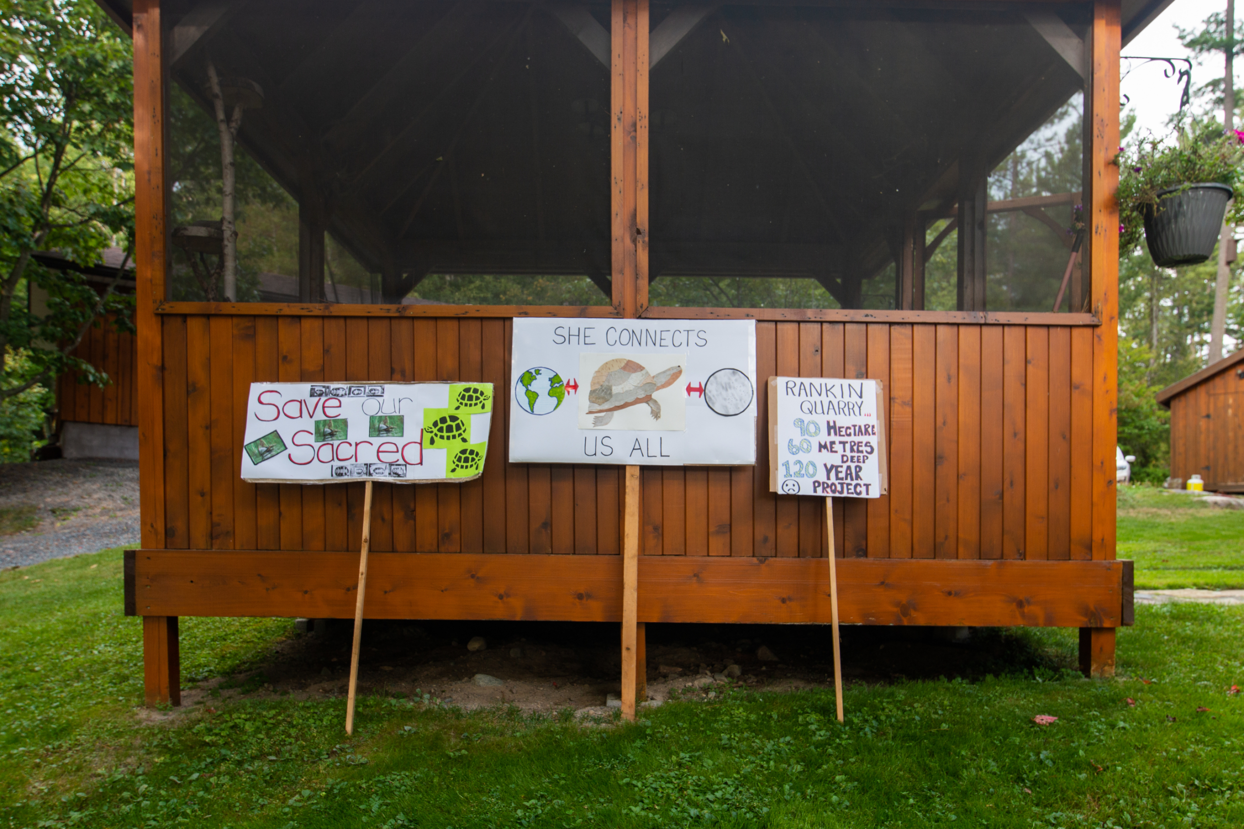 Signs reading "save our sacred turrtles," "she connects us all," and "rankin quarry, 90 hectares, 60 metres deep, 120 year project" with drawings of turtles and the earth, leaning against a gazebo