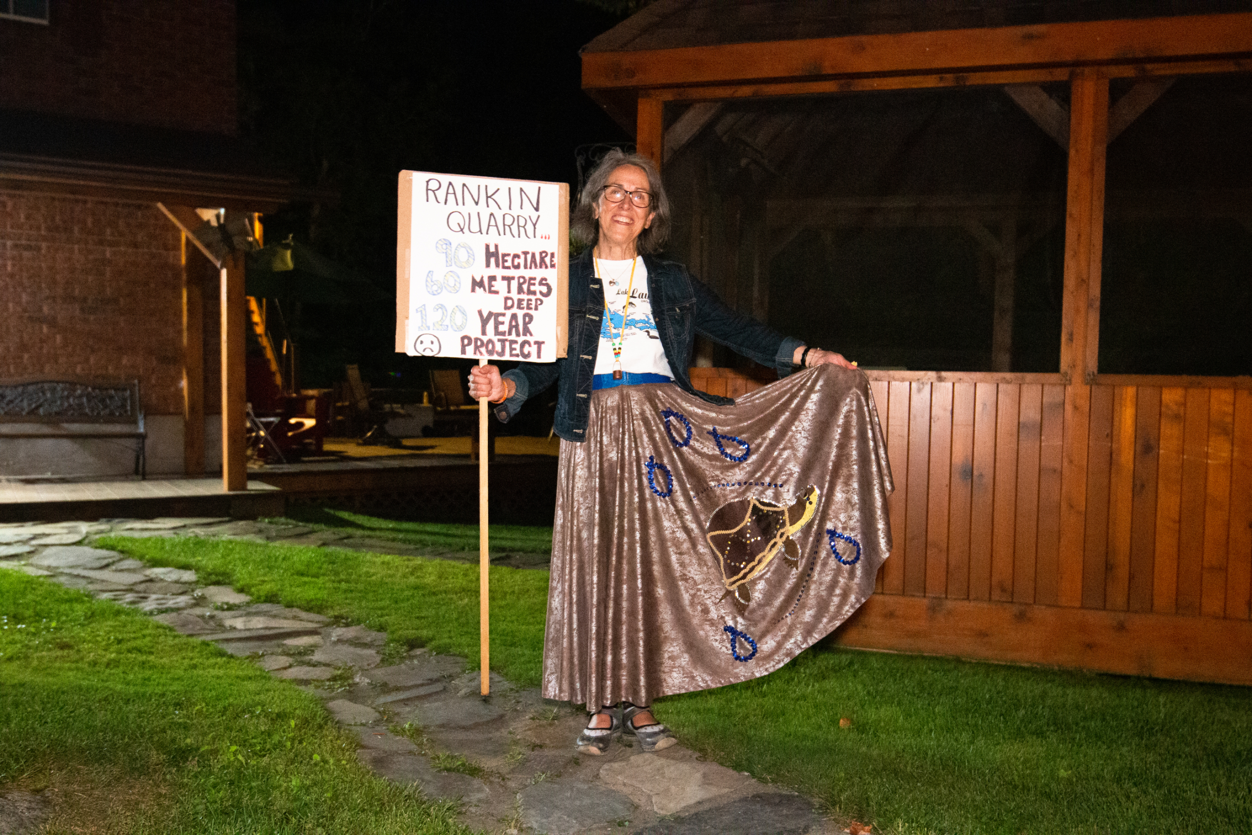 Janice Gamble, wearing a turtle skirt, poses with a sign reading "Rankin quarry, 90 hgectares, 60 metres deep, 120 year project"