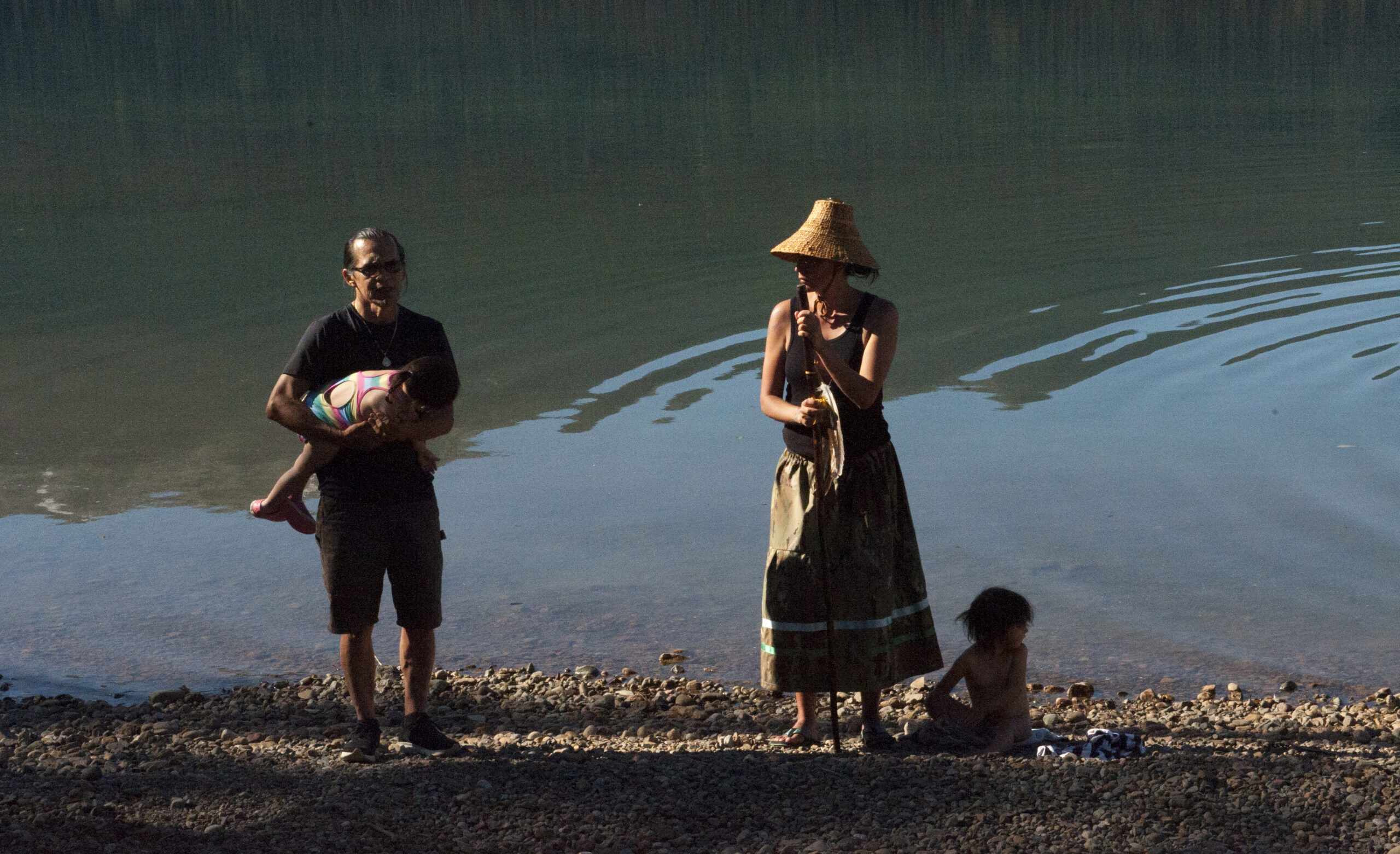 Wet'suwet'en Hereditary Chief Woos and Sleydo' Molly Wickham stand on the shore of Wedzin Bin (Morice Lake). Woos holds one of his children, while Wickham's daughter sits at her feet