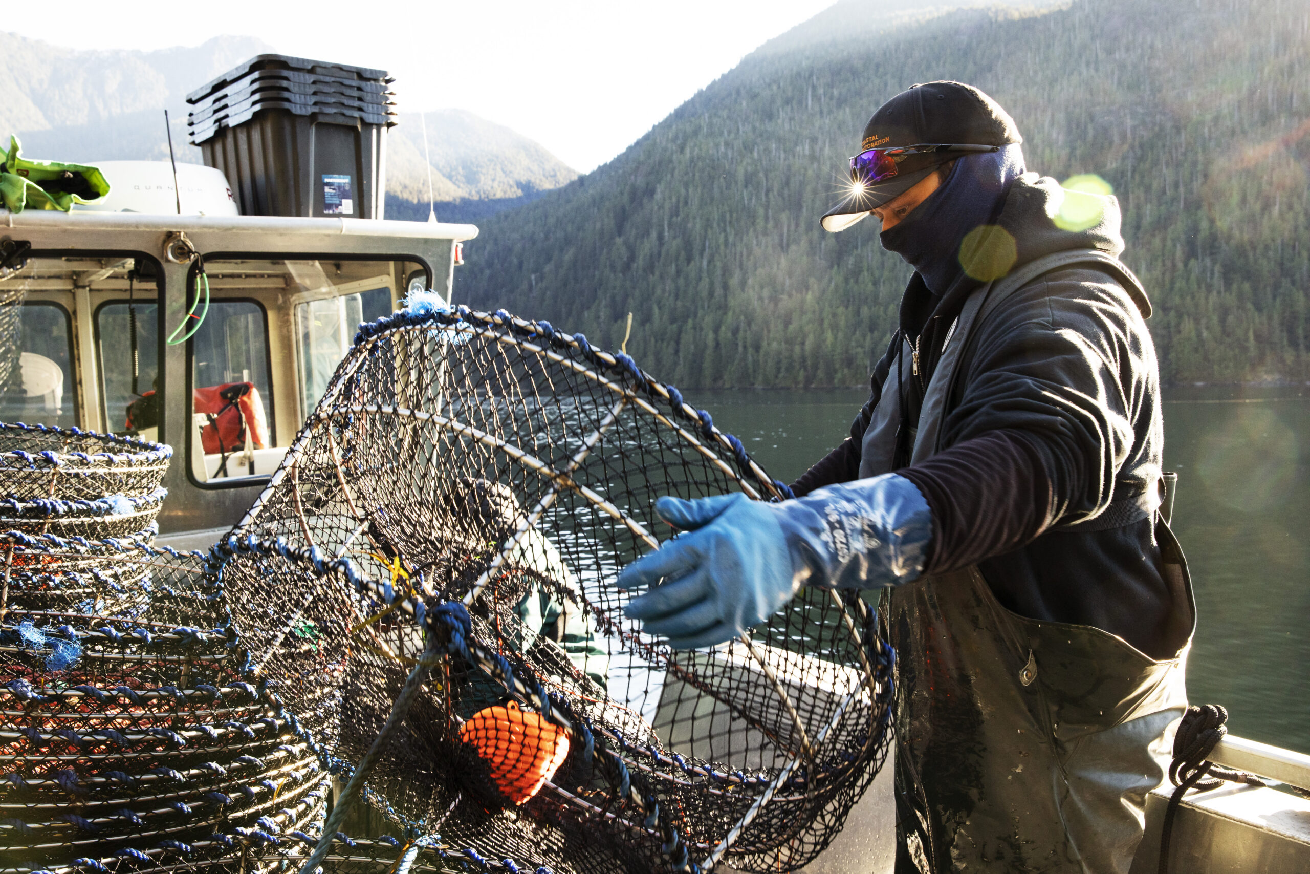 Coastal Restoration Society technician James George, wears a ball cap, overalls and blue gloves. He moving a prawns trap used to capture European green crabs aboard a boat in Bedwell Sound.