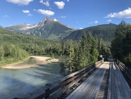 a wood bridge crosses the North Thompson River, in the background logging cutblocks are visible on the moutains