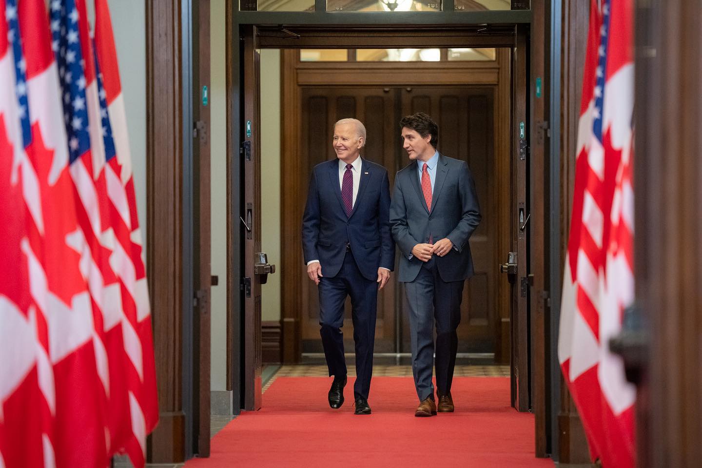 Joe Biden, wearing a dark blue suit, walks down a red carpet lined with U.S. and Canadian flags next to Justin Trudeau in a grey suit.