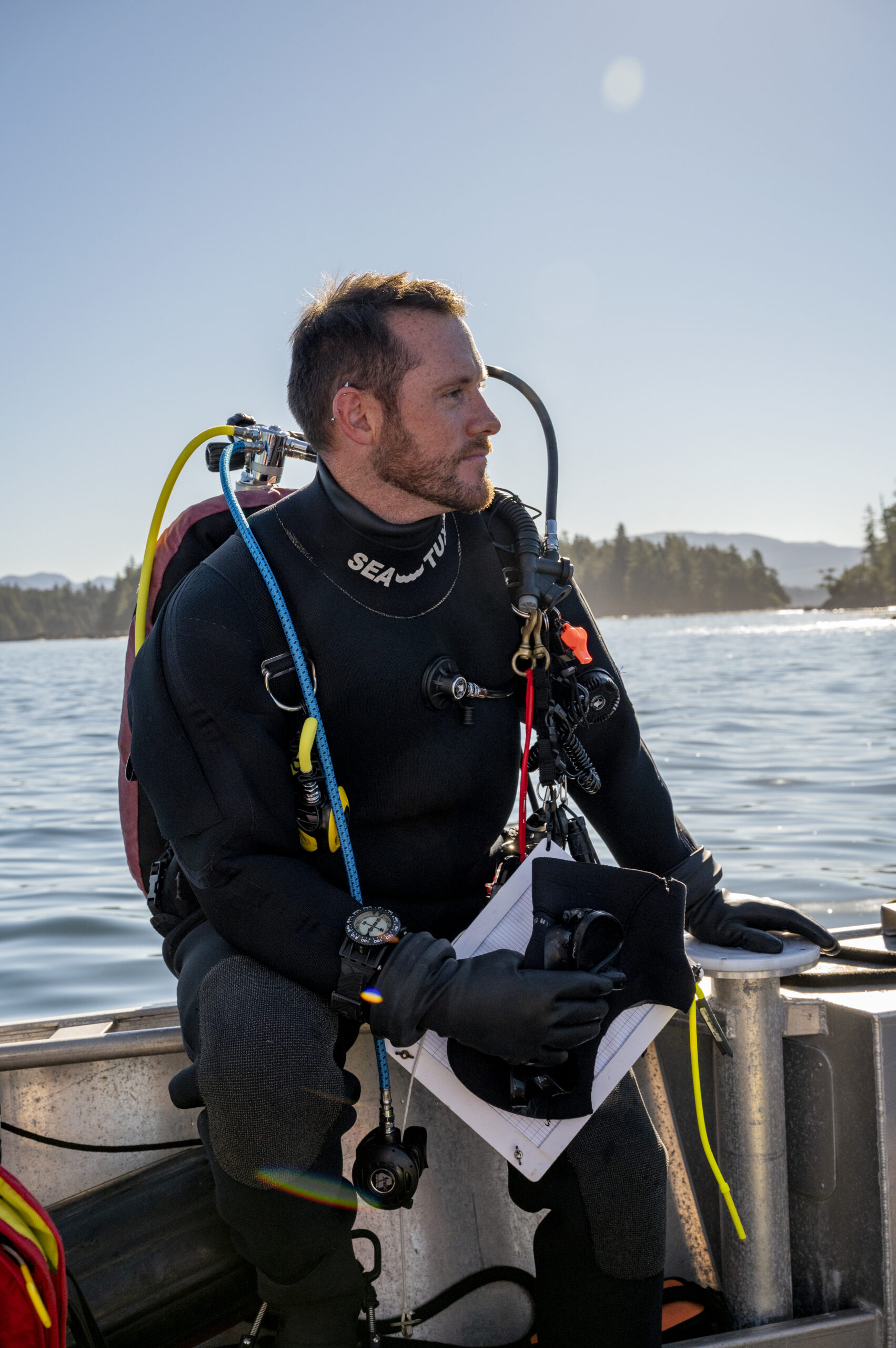 A portrait of marine ecologist Kieran Cox in his dry suit, sitting on the side of his research boat with the ocean and trees in the background