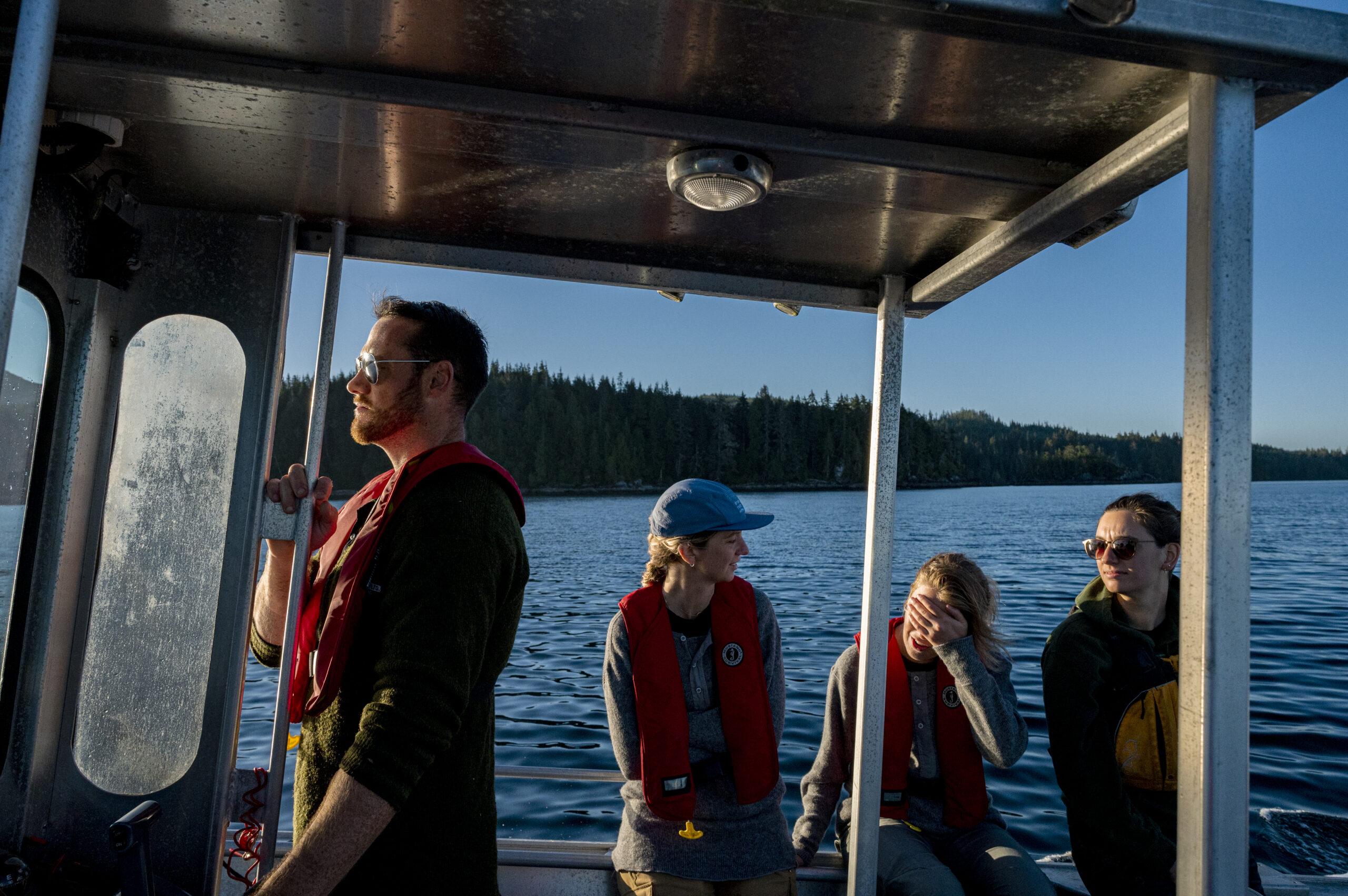 Marine ecologist Kieran Cox drives the research boat as his colleagues Bridget Maher, Claire Attridge and Em Lim lean against the side of the boat. The ocean and forested land are seen in the background.