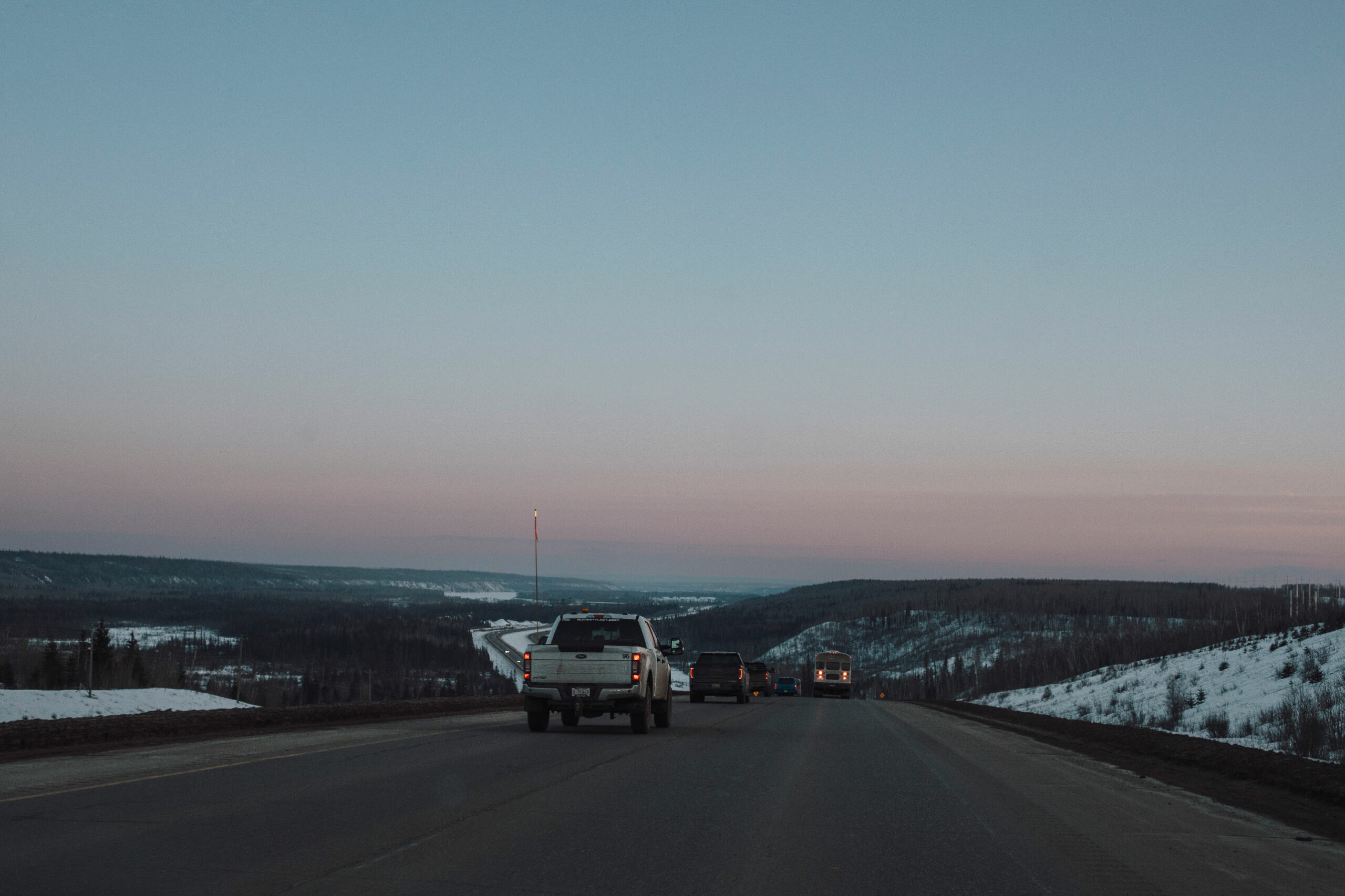 Pickup trucks drive down a wide highway under a twilight sky.
