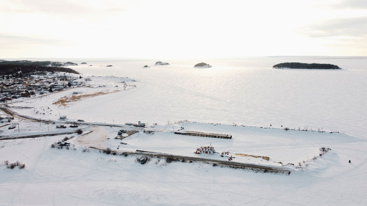 A snow-covered barge at a port on frozen Lake Athabasca with the community of Fort Chipewyan visible on the shore.