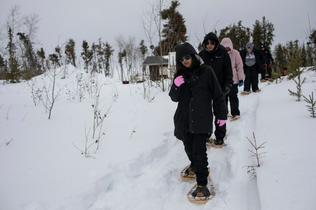 Young people snowshoe on a snowy path with conifers in the background