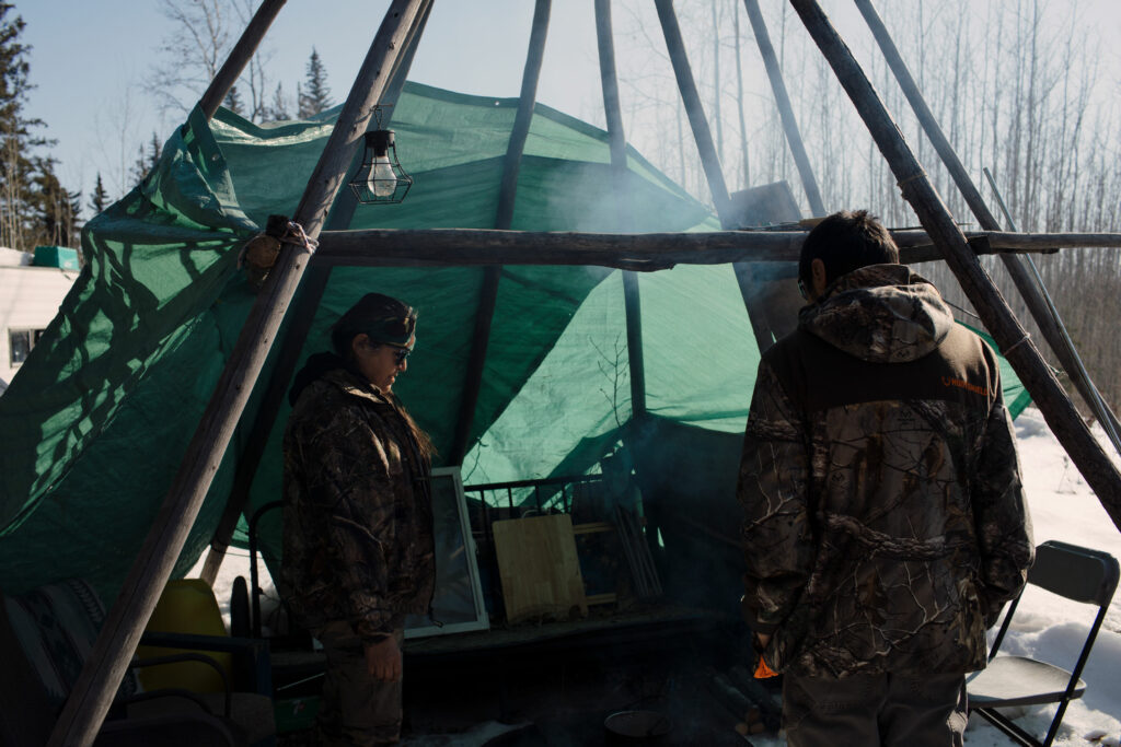 People in camouflaged jackets stand under a teepee on a wintry day