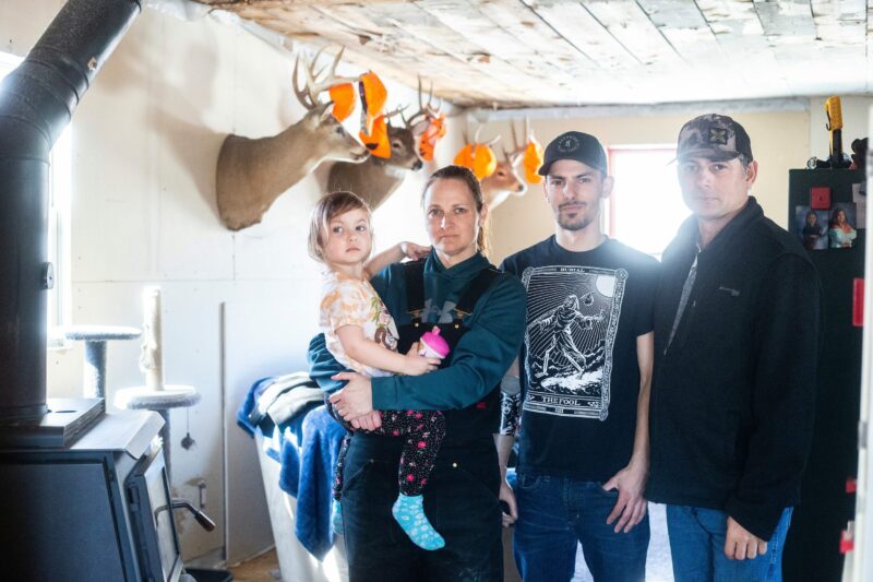 Georgina, holding her daughter Callie, stands next to Wesley and Josh Mustard in front of mounted deer heads with orange caps hanging from the antlers in the living room of their Vivian, Manitoba home