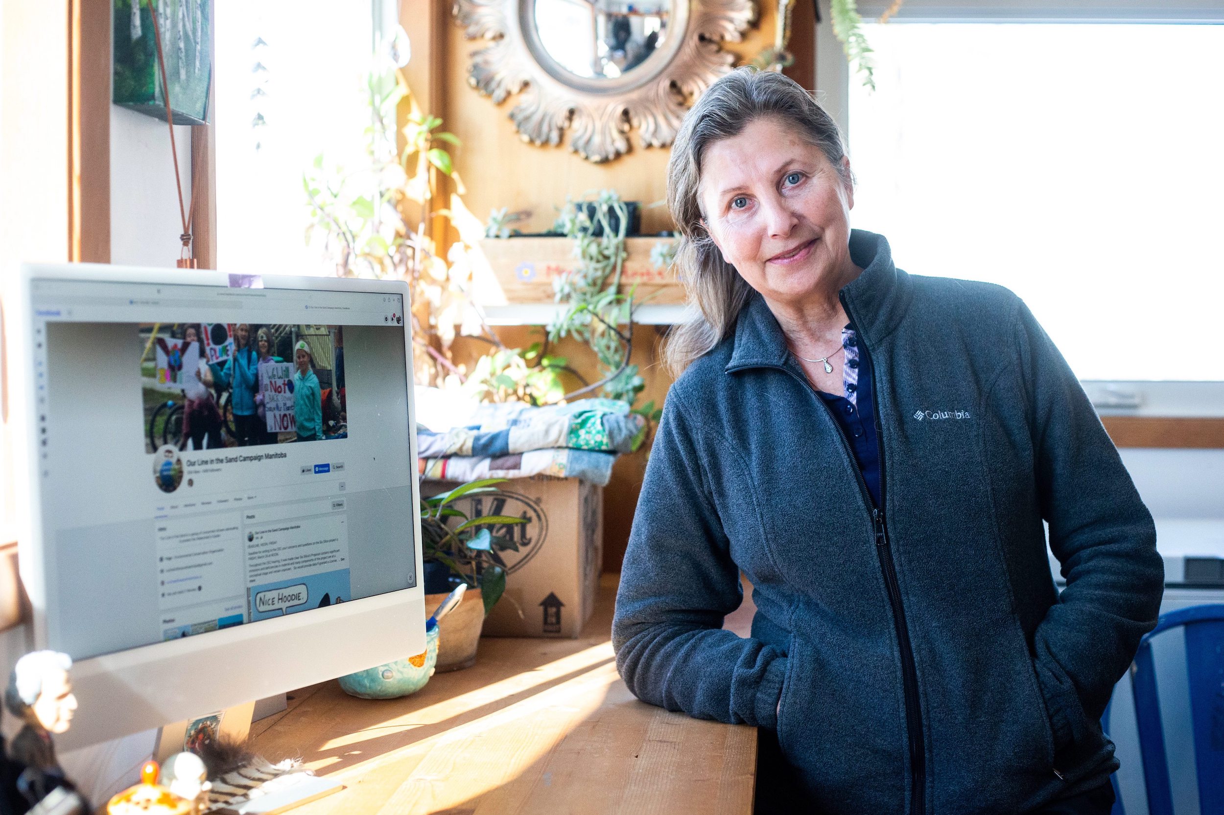 Tangi Bell, wearing a grey sweater, stands in her home office next to a computer displaying the Our Line in the Sand facebook page