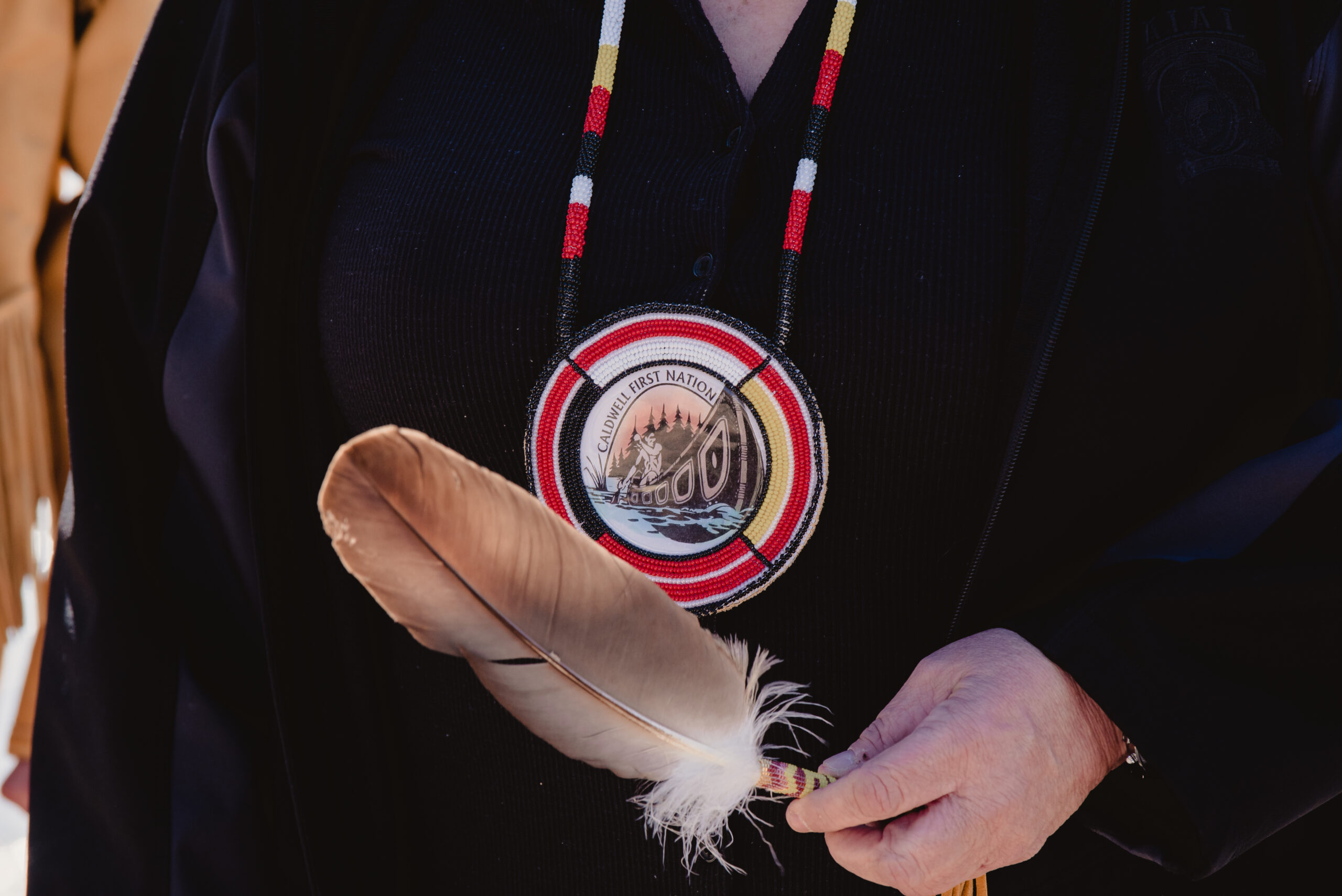 A woman holds an eagle feather in her hand, in front of her black shirt and a Caldwell First Nation badge.