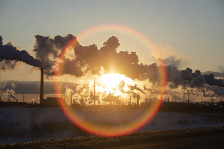 The sun setting behind an industrial plant, which is emitting large plumes into the sky.
