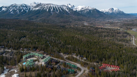 An aerial photograph shows a strech of forest below snow dusted peaks with a residential development in the foreground