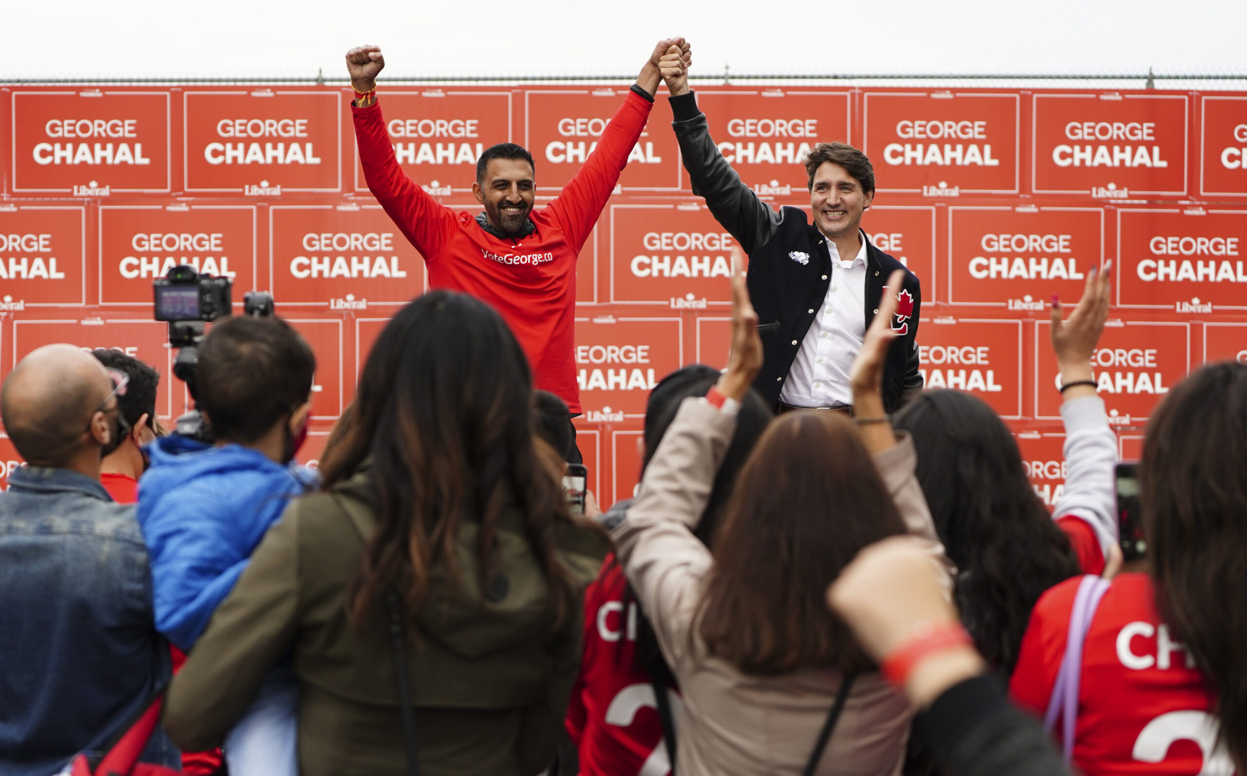 A man in red raises his hands and holds the raised hand of another man standing in front of a crowd with Liberal Party red signs in the background.