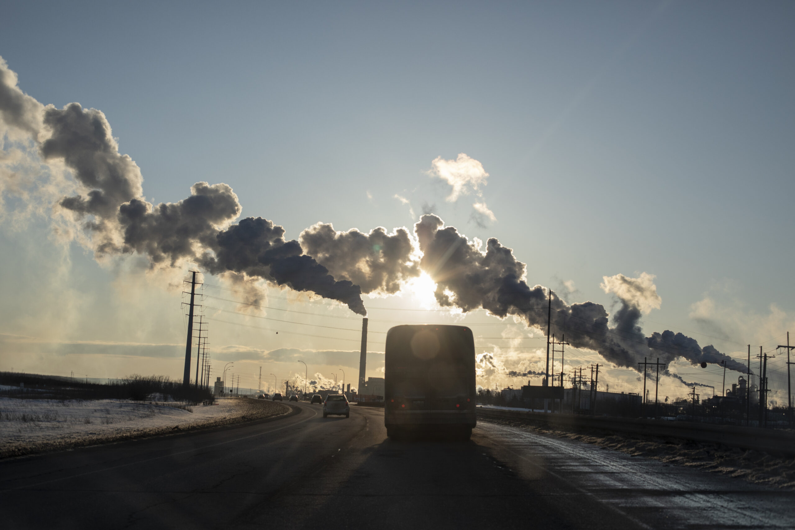 A bus drives down the road with industrial facilities in the background emitting smoke or vapour.