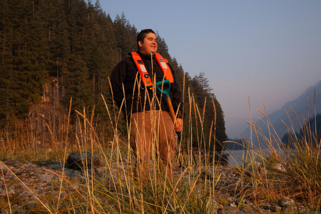 A portrait of Travis George with some grasses in the foreground and trees in the background. Travis is wearing an orange life vest and holding a pitchfork used to harvest clams from the sand.
