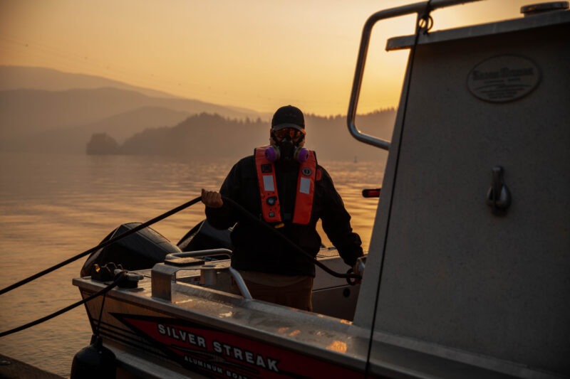 Travis George, wearing a mask and orange life vest, unmoors the boat. The smoky haze has given the area an orange glow