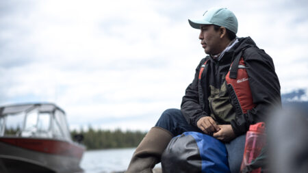 Ryan Kyikavichik in Yukon looks out on the water, wearing a vest boot and hat with a boat in the background on a cloudy day