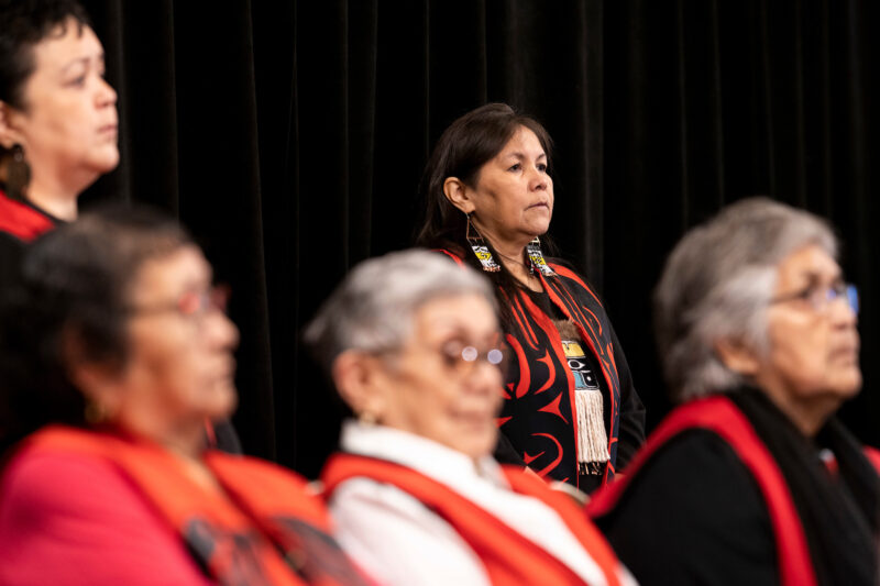 A woman stands in Indigenous regalia before a black curtain