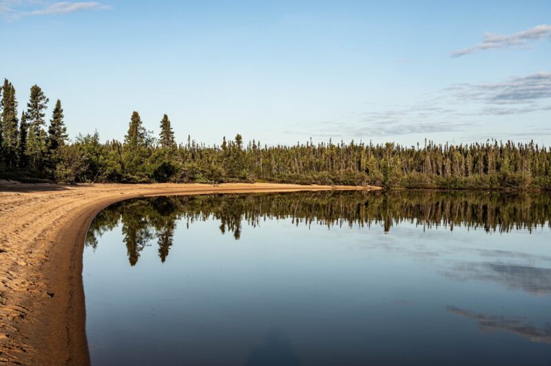 A northern Manitoba lake shoreline surrounded by evergreen trees. The treeline is reflected on the water