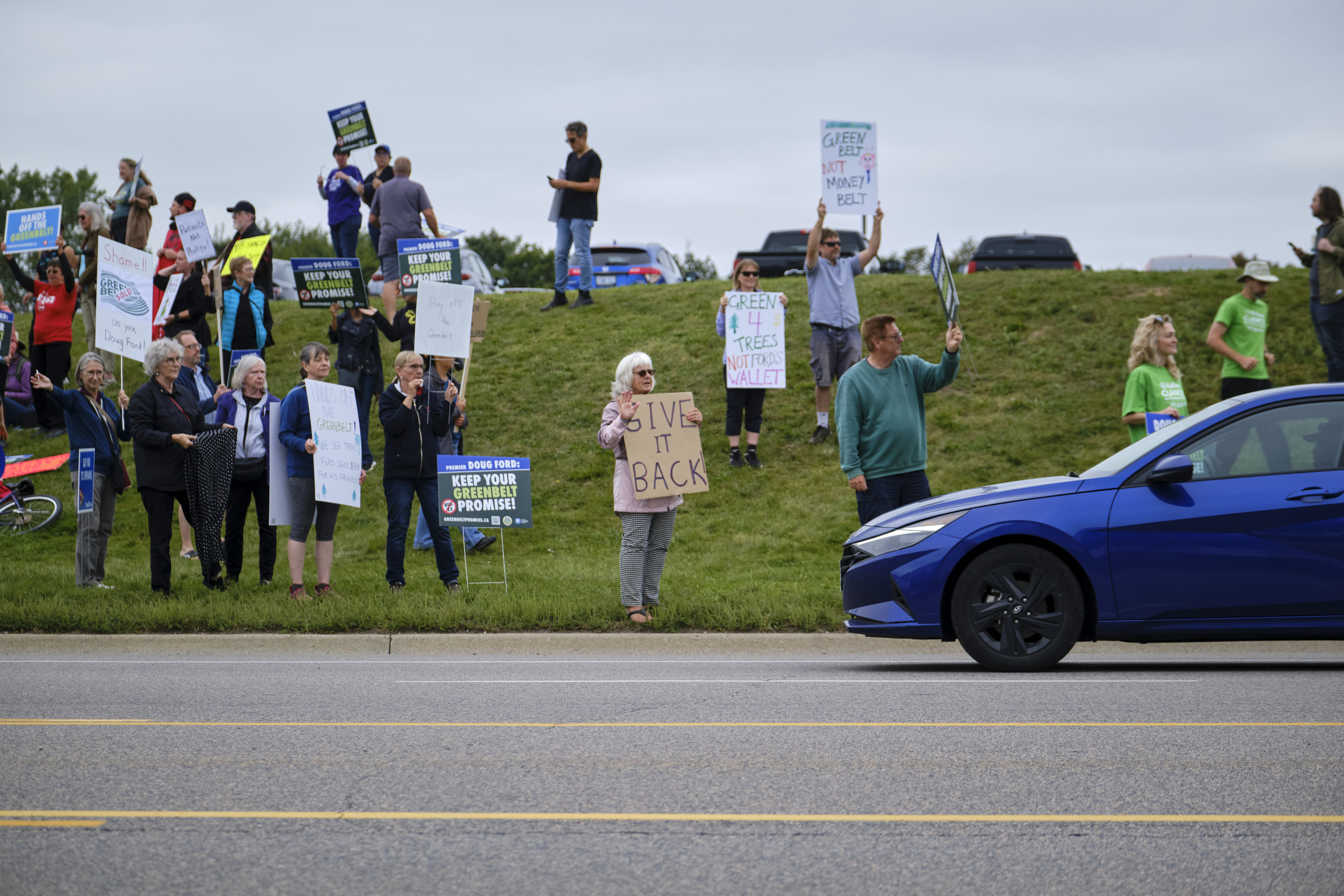 Citizens by the side of the road protest the Ontario government's Greenbelt changes with signs saying "Give it back" and "Keep your Greenbelt promise"