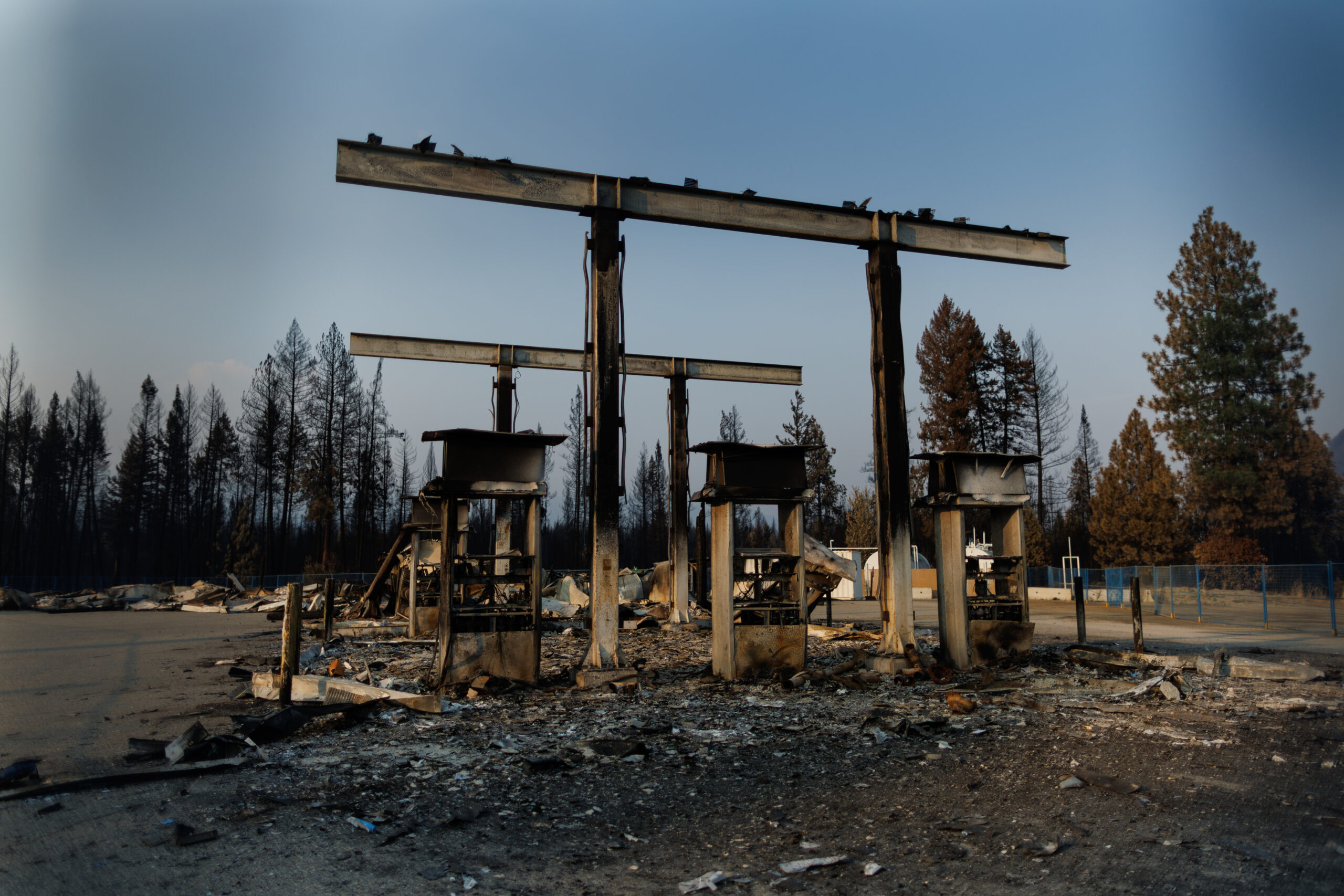 A gas station is burnt black in the Squilax (Little Shuswap) community east of Kamloops, B.C. The ground and the trees in the background are blackened, and the sky is blue but hazy above.