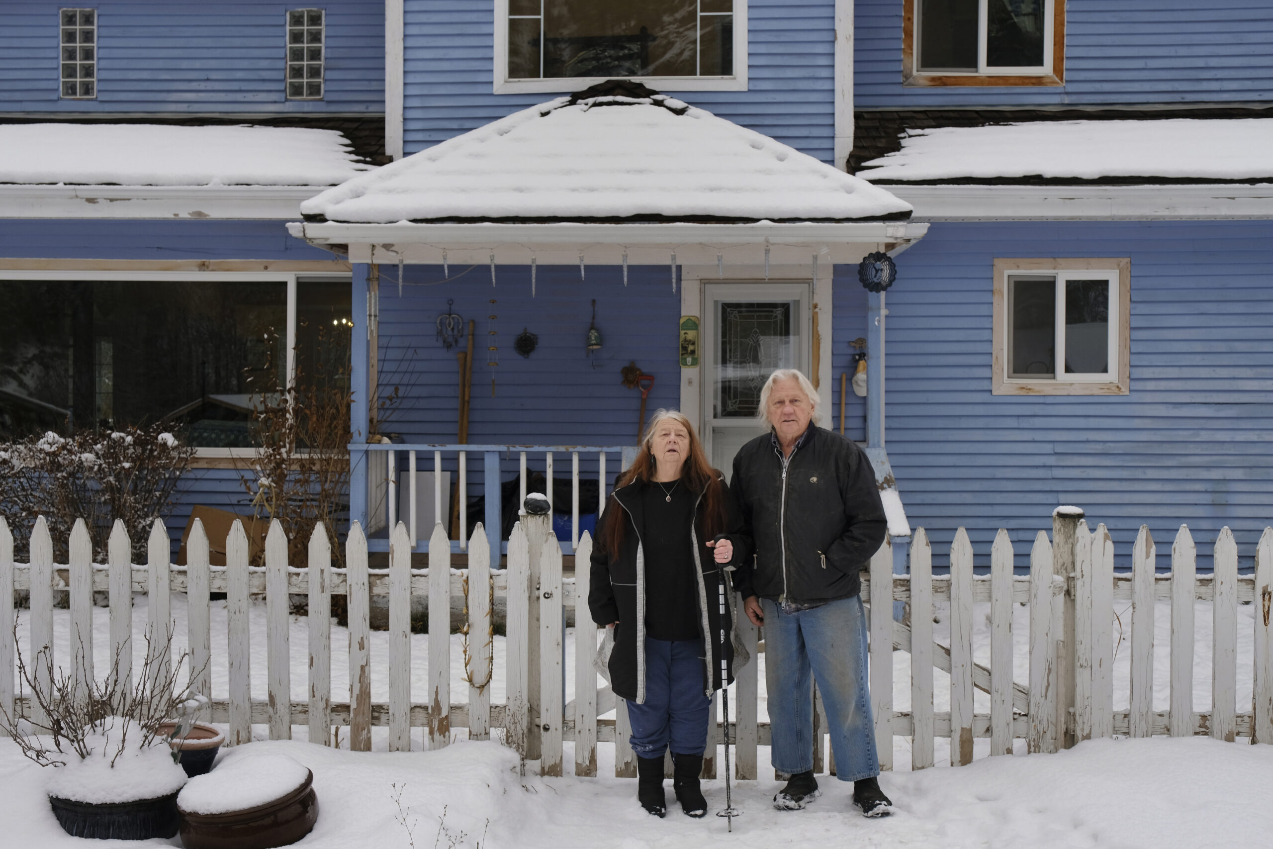 A brother and sister stand in front of their blue home. The ground is covered in snow and they are in front of a white picket fence.