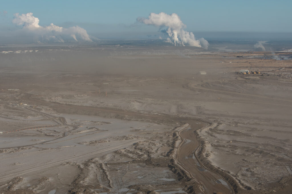 Plumes rise on the distant horizon where upgraders from Suncor Base plant and the Syncrude Mldred Lake plant are visible, with open mines in the foreground