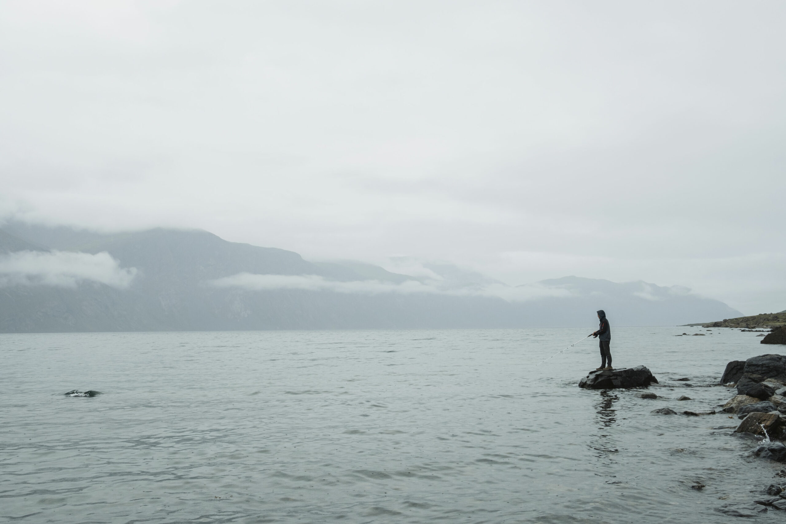 A participant from the Youth Leadership Program fishes for Arctic char in Torngat Mountains National Park. They stand on a rock, with cloudy landscape and waters behind them.