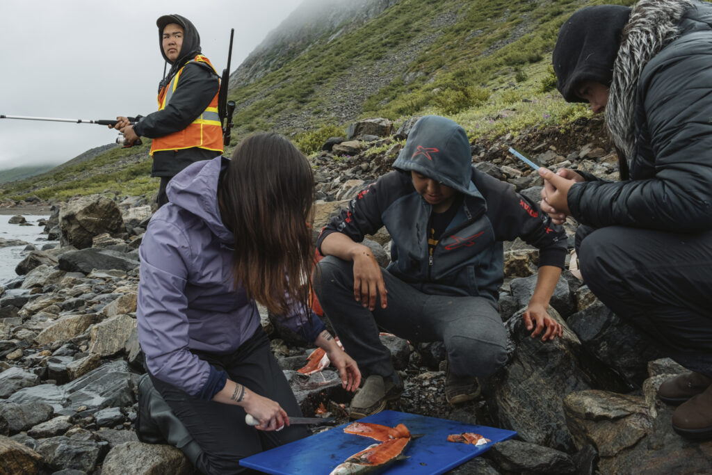 People fillet a fresh Arctic char on a cutting board at the shore with mist obscuring the mountainside behind them