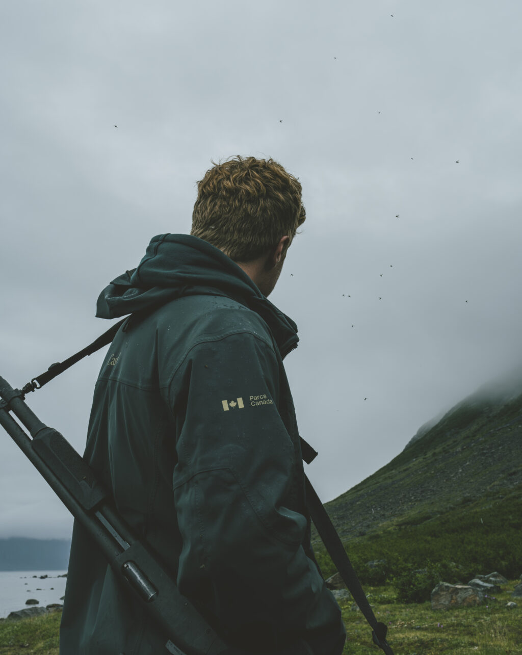 A man in a Parks Canada jacket looks away from the camera with gun slung over his shoulder and bugs flying around his head