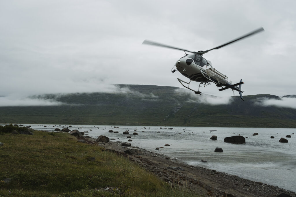 A helicopter lands at the rocky shore of Nachvak Fiord with mist in the background